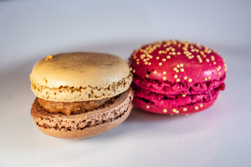 Colorful macarons on white floral plate against warm textured background. Red, beige, and chocolate tones with visible fillings. Cozy and inviting dessert still life.