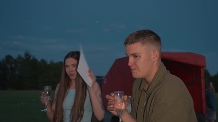 man sipping champagne from glass while woman waves certificate to swat buzzing insects around them on grassy field at dusk creating playful protective moment after scenic hot air balloon flight - Powered by Adobe