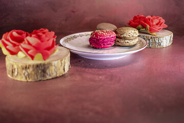 Assorted macarons in red, brown, and beige on decorative white plate. Red macaron with sprinkles stands out. Wooden coaster and red rose add warmth and cozy, intimate atmosphere.