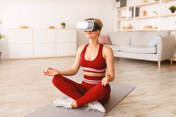 Modern Technologies And Sports. Calm peaceful woman wearing VR headset sitting in lotus pose, meditating and practicing yoga indoors at home. Lady using virtual reality glasses for home practice
