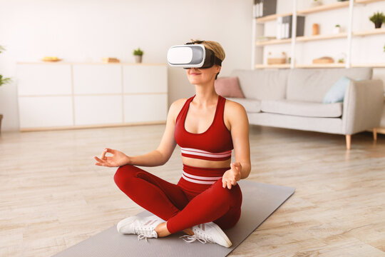 Modern Technologies And Sports. Calm peaceful woman wearing VR headset sitting in lotus pose, meditating and practicing yoga indoors at home. Lady using virtual reality glasses for home practice