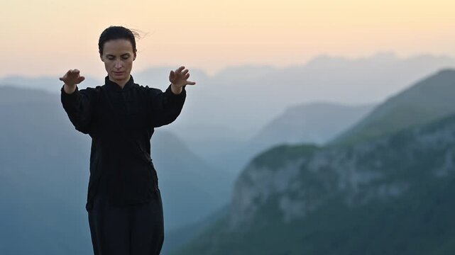 Woman practicing Tai Chi in traditional black outfit on mountain at sunrise, arms outstretched in meditation pose, focusing on balance, energy, and mindfulness in nature.