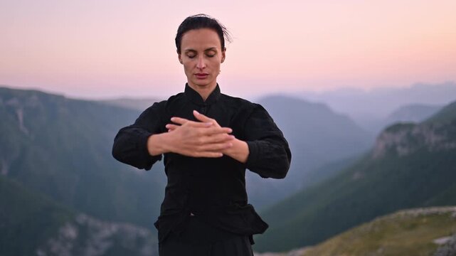Woman practicing Tai Chi in traditional black outfit on mountain at sunrise, arms outstretched in meditation pose, focusing on balance, energy, and mindfulness in nature.