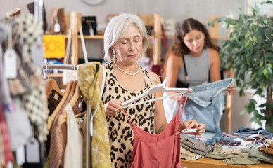Hesitant old-aged female customer reviewing summer dresses during shopping in clothing store