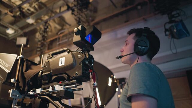 Cameraman in headset operating professional television camera inside broadcast studio while recording presenter on blue screen stage with teleprompter, lighting gear, ceiling rig, and video monitor in