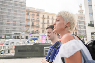 Fototapeta premium A man and a woman are sitting on a bench in a city. The woman is wearing a white shirt and a backpack. The scene is set in a city with a lot of buildings