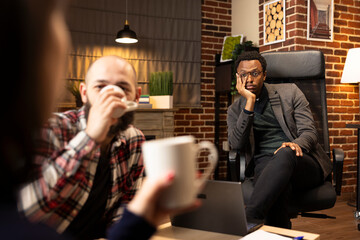 Black therapist seated on chair, leaning on hand and attentively observing caucasian couple with cups. White man sipping tea as wife speaks during emotional counseling session in private office.