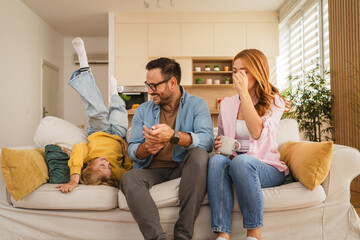 Family relaxing on sofa: daughter playing while parents using smart phone and drinking coffee