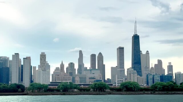 A view of Chicago's skyline from Lake Michigan, with a row of trees in front of the city's iconic skyscrapers under a cloudy sky.