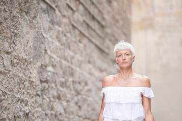 A woman in a white dress stands in front of a brick wall. She looks sad and is wearing earrings