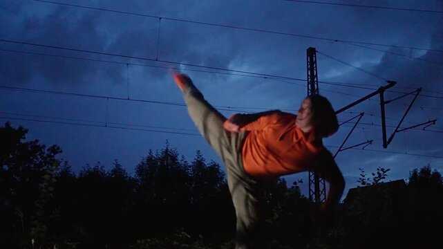 An athlete, a martial artist, performs a kick, against the backdrop of the evening sky and against the backdrop of a passing train, a cinematic moment in super slow motion