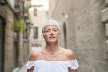 A woman with white hair and a white dress stands in front of a brick wall. She has a serious expression on her face