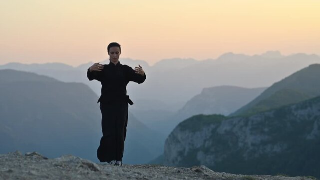 Woman practicing Tai Chi in traditional black outfit on mountain at sunrise, arms outstretched in meditation pose, focusing on balance, energy, and mindfulness in nature.