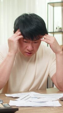 Asian man sitting at home table grips head, unpaid bills scattered beside calculator and laptop. Chinese guy sighs deeply, showing heavy financial stress and urgent need to balance household budget.