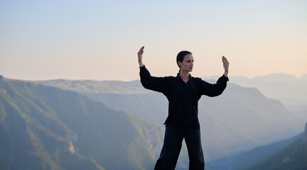 Woman practicing Tai Chi in traditional black outfit on mountain at sunrise, arms outstretched in...