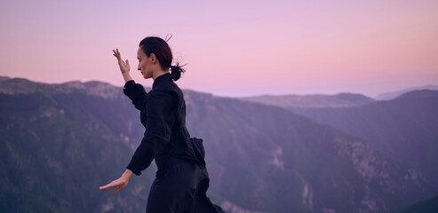 Woman practicing Tai Chi in traditional black outfit on mountain at sunrise, arms outstretched in...