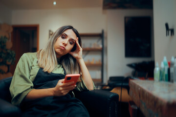 Bored Hairdresser Checking her Phone Having No Clients. Unhappy tired hairstylist sitting on the...