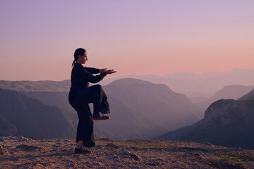 Woman practicing Tai Chi in traditional black outfit on mountain at sunrise, arms outstretched in meditation pose focusing on balance, energy, and mindfulness in nature.
