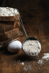 Baking Still Life. A bag of flour with a sifter full of flour leaning on the bag, an egg and whisk found out the scene on a rustic wood table.