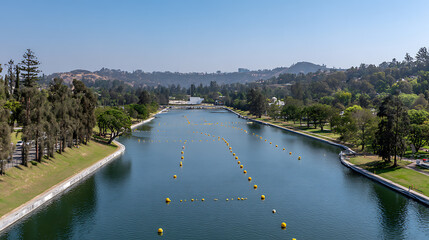 Aerial view of a calm lake with floating buoys in a park surrounded by trees and hills under clear blue sky