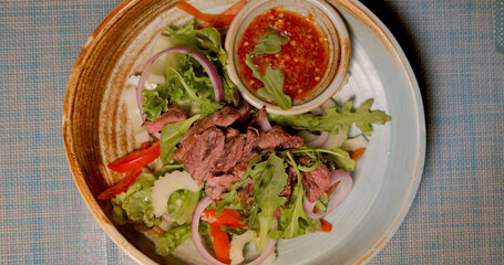 Grilled beef salad with red onion, arugula, and spicy dipping sauce on rustic ceramic plate, overhead view. Fresh ingredients, vibrant colors, healthy Thai cuisine concept.