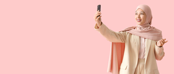 Young Muslim businesswoman with mobile phone video chatting on pink background