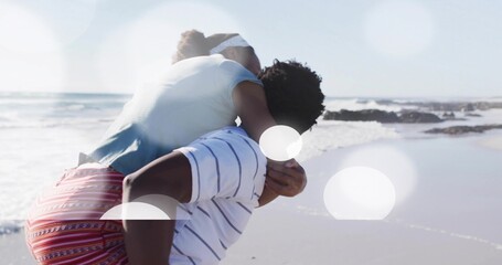Man carrying woman piggyback along sandy beach under bright sunlight barefoot, with gentle waves