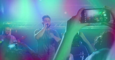 Singer singing into mic at venue under colorful truss lights, with guitarist smartphones recording