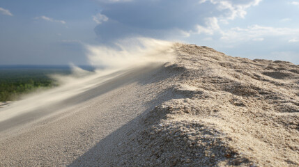 Sandy dune with fine grains of sand being blown by wind under cloudy sky, creating dynamic and serene landscape. scene captures beauty of nature and movement of sand