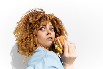 Surprised young woman with curly hair holding a hamburger, expressing shock against a white background, conveying a humorous take on indulgence and fast food