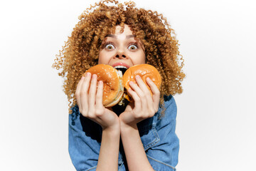 Happy young woman with curly hair joyfully holding two burgers, promoting healthy eating and fun food choices with a vibrant, playful expression