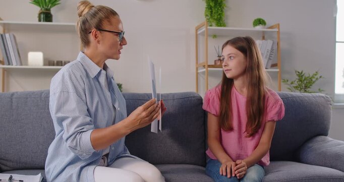 Female therapist and child on sofa discuss emotions with cards during a counseling session in calm office. They explore feelings and build trust. Psychology assessment supports communication.