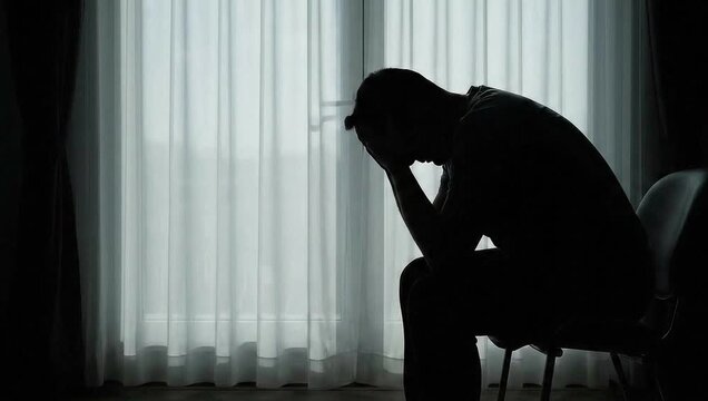 Silhouette of a person in deep thought sitting alone by a window in a dimly lit room during the evening
