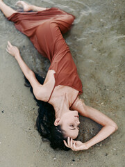 A graceful woman lies on a sandy beach wearing a rust dress, sunlit skin and relaxed pose, capturing calm elegance, texture, and natural mood.