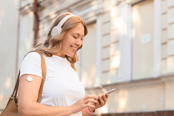 Diabetic woman in headphones with glucose sensor using mobile phone on street