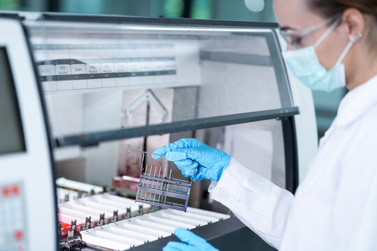 A female scientist in a lab coat and mask loads a rack of test tubes into a large medical testing machine. She is carefully preparing samples for important health analysis. - Powered by Adobe