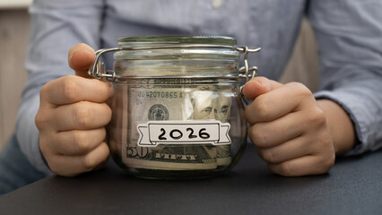 Unrecognizable woman holding Saving Money In Glass Jar filled with Dollars banknotes. 2026 year transcription in front of jar. Managing personal finances extra income for future insecurity background