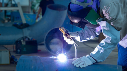 A welder focuses on joining metal pipes in a factory. Bright sparks fly in the air as he uses a...