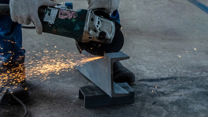 At a construction site, a worker uses an angle grinder to cut a steel beam, creating sparks that...