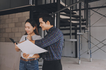 Construction manager and engineer dressed in orange work vests and hard helmets explore construction documentation on the building site near the steel frames