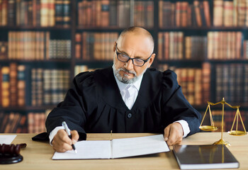 Judge in robe signing legal document in courtroom. Professional old magistrate sitting at wooden...