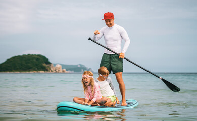 Dad paddles while kids sit in front of him on stand-up paddleboard, having fun on calm ocean waves. Family activity, connection, and laughter in motion.