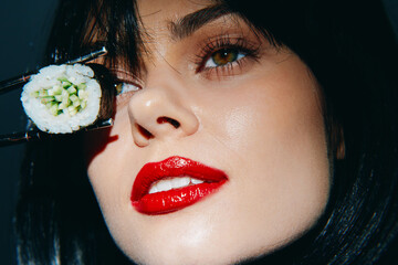 Close-up of a model with dark hair holding sushi with chopsticks, featuring striking red lips and an expressive gaze, highlighting Japanese cuisine.