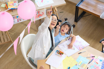 A teacher points upward while drawing on a paper with children. They are engaged in an art lesson together, smiling and having fun