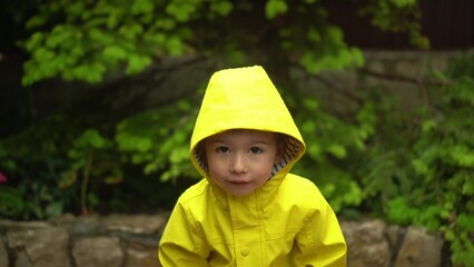 child in yellow raincoat with hood stands in the rain smiling and waving hands looking at the camera with green garden background