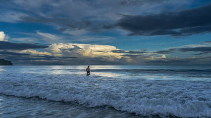 Early morning. The ocean waves are rolling towards the shore, foaming. Glare on the surface. The silhouette of a man knee-deep in water, walking into the sea. The clouds in the sky are highlighted  