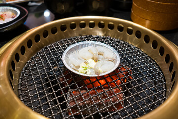 A small aluminum dish of raw scallops and shrimp cooks on a hot charcoal grill at a Korean BBQ restaurant