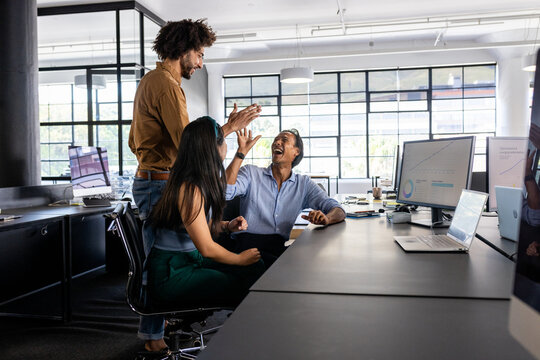 Business team celebrating success at office desk with high fives and laughter - Powered by Adobe