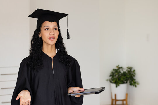 Graduate in cap and gown holding tablet, speaking confidently at home - Powered by Adobe