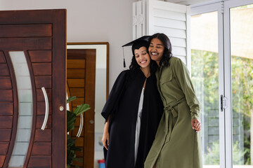 Proud graduate in cap and gown hugging supportive friend at home doorway